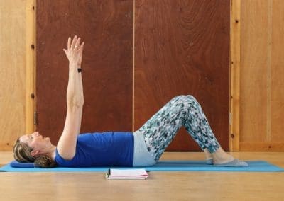 a woman lying on her back on a blue mat, participating in a Lucy Purrier Physio Pilates class. She is in a calm setting with wooden doors in the background, indicative of a community hall. The woman, wearing a blue Pilates shirt and patterned leggings, has her arms extended straight upward, focusing intently on her exercise. A notebook beside her on the mat suggests a structured approach to tracking her Pilates regimen. This serene scene captures the essence of focused physical wellness activity in a supportive group setting.