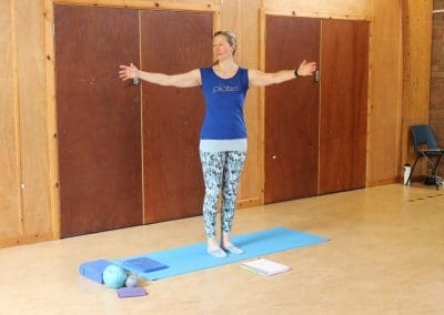 a focused moment in a Lucy Purrier Physio Pilates class, held in a spacious hall with wooden interiors. A middle-aged woman in a blue Pilates shirt and patterned leggings stands on a blue mat, her arms extended wide in a balancing exercise. Essential Pilates equipment, such as a foam roller and a couple of textured massage balls, lies beside her on the mat. A notepad rests on the floor, likely used for tracking progress or exercises, hinting at a structured session. The background shows wooden doors and a chair, adding to the community hall ambiance.