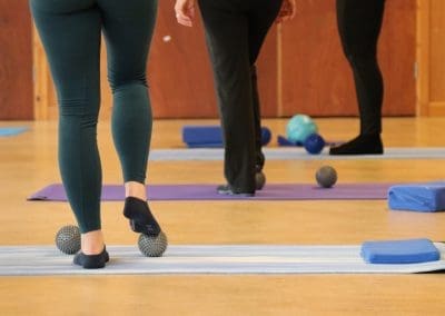 captures a close-up view of participants' legs and feet during a Lucy Purrier Physio Pilates class, illustrating a focus on foot and ankle exercises. Two women are seen standing on textured massage balls, enhancing their balance and stimulating the soles of their feet. The one in the foreground wears dark green leggings and the other in black, both engaging in this therapeutic exercise. Scattered around are Pilates mats and equipment, including blue and green balls and foam blocks, set against the backdrop of a well-lit wooden hall. This scene highlights the attention to detail and physical therapy aspects integrated into the Lucy Purrier Physio Pilates program.