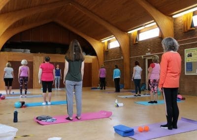 group fitness class at St. Peter's Church hall, participating in a Lucy Purrier Physio Pilates session. Women of various ages stand on colorful mats in rows, engaging in light stretching exercises. The wooden arched ceiling and brick walls of the hall provide a warm, inviting atmosphere. Some participants are in motion, adjusting their positions, while others are attentively listening to instructions. Physiotherapy equipment like small balls and foam rollers are scattered across the mats, indicating a focus on therapeutic exercises.