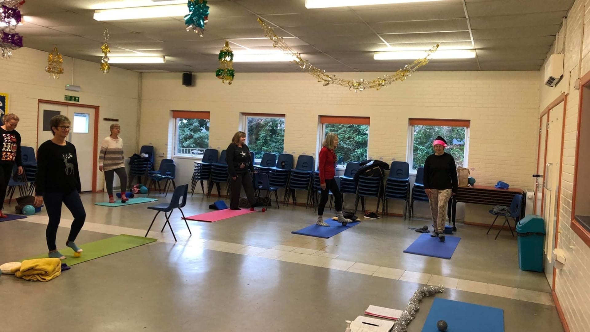 A group of participants in a pilates class in Broadstone, exercising in a spacious community room. The room is decorated with festive hanging ornaments, and each participant is using a yoga mat for exercises. The environment is casual and friendly, with a few props like resistance bands and small exercise balls visible. The pilates Broadstone class setting emphasizes a supportive and relaxed atmosphere for people of varying fitness levels.