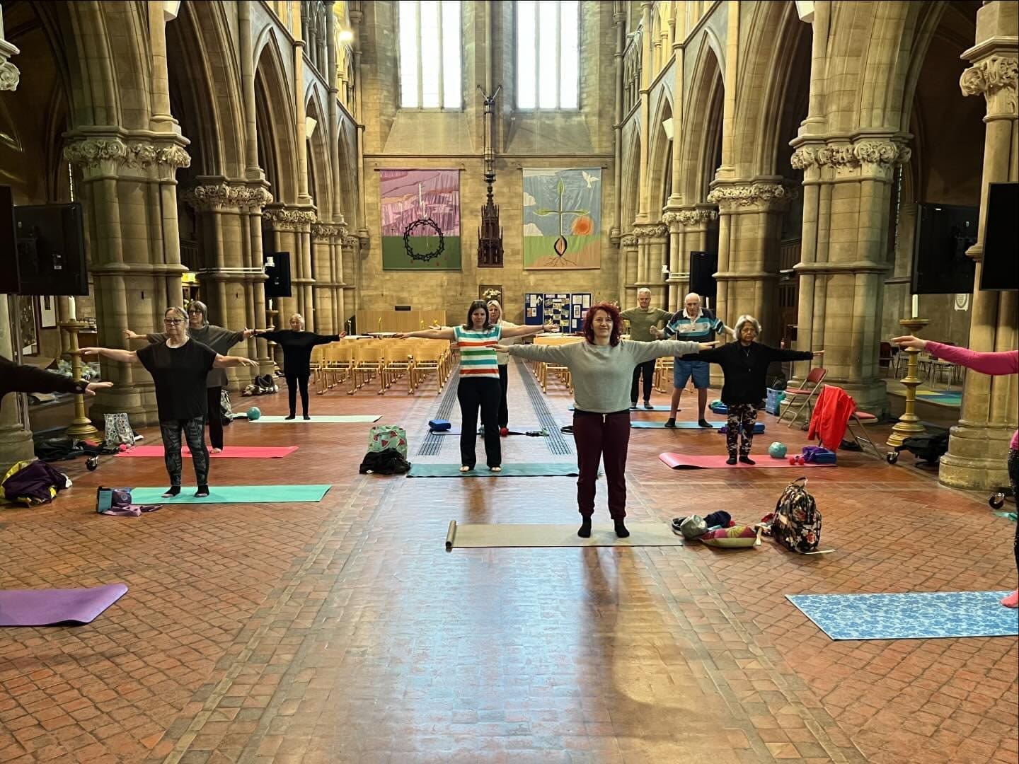 A pilates class taking place in a unique setting that resembles a historic church or cathedral, with participants spread out on yoga mats. The pilates studio atmosphere is characterized by high vaulted ceilings, large stone pillars, and colorful banners hanging on the walls. Several people are performing stretching exercises in a spacious, open area, with mats, resistance bands, and small exercise balls visible. The scene captures a welcoming and supportive group exercise environment.