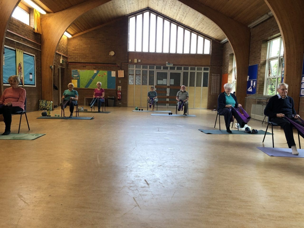 A wide-angle view of a senior physio Pilates class, with participants seated on chairs spaced across a large hall, each holding a purple stretch band. The elderly participants, dressed in comfortable attire, engage in gentle exercises, guided by their bands and the peaceful, well-lit environment of the community hall. The architectural details, such as the wooden beams and large windows, contribute to the serene atmosphere of the session.