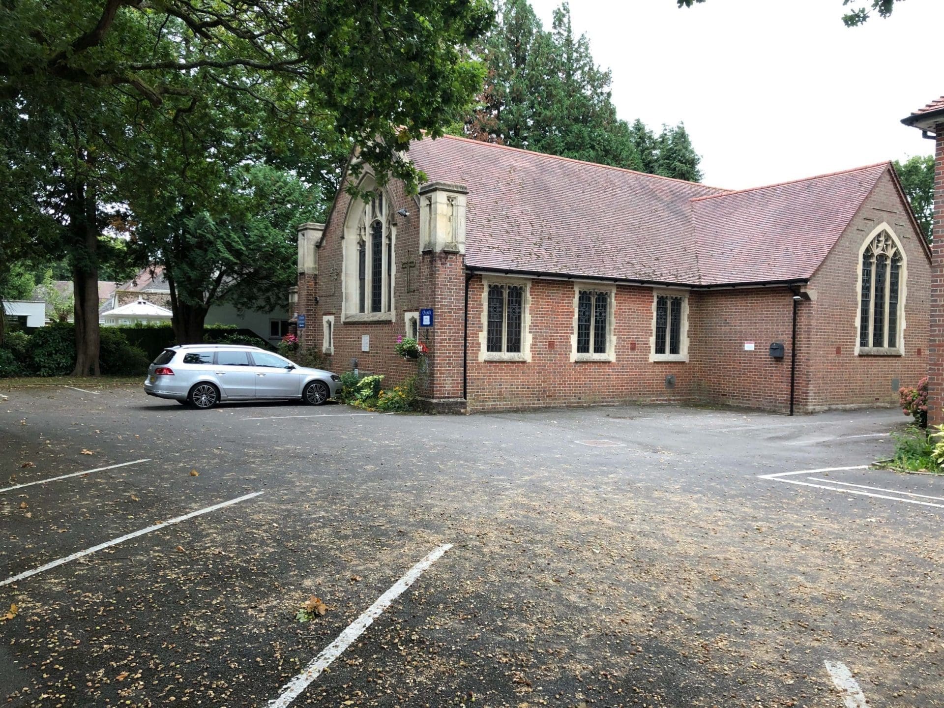The exterior view of the United Reformed Church Hall in Broadstone, a venue for Pilates classes. This one-story building, constructed with red brick and featuring pointed Gothic windows, is surrounded by lush green trees. The parking area in front of the hall, marked with faded lines, hosts a silver car, emphasising the hall's accessibility and community use.